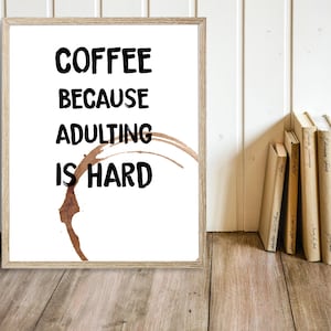 Puede incluir: Una impresión enmarcada blanca con el texto "COFFEE BECAUSE ADULTING IS HARD" en letras negras. Una mancha de café marrón es visible en la impresión.
