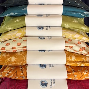 May include: A stack of rectangular fabric eye pillows featuring various patterns including floral, celestial, bee, snail, sun, and abstract designs. Each pillow is wrapped in a white paper band labeled Flaxseed Tranquility with a circular logo.