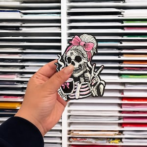 May include: An embroidered patch featuring a skeleton with a high bun hairstyle, a pink bow, and a striped shirt. The skeleton is holding a small jar with a pink bow and making a peace sign gesture with one hand, rendered in black, white, and pink.
