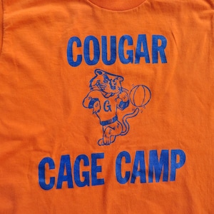Puede incluir: Camiseta naranja con un gr&aacute;fico azul de un puma de dibujos animados sosteniendo una pelota de baloncesto. El texto "COUGAR CAGE CAMP" est&aacute; impreso en azul debajo del gr&aacute;fico.