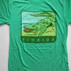 May include: Green t-shirt featuring a vintage-style illustration of an alligator with the word "FLORIDA" printed below it.