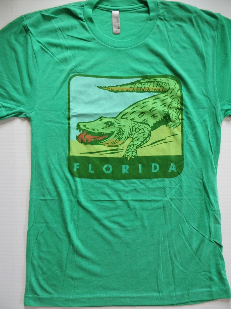 May include: Green t-shirt featuring a vintage-style illustration of an alligator with the word "FLORIDA" printed below it.