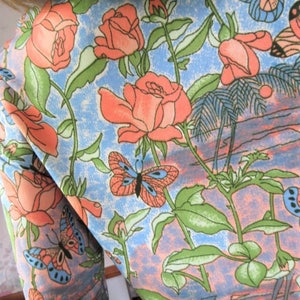 K&ouml;nnte beinhalten: Ein blauer Stoff mit einem Blumenmuster mit rosa Rosen, gr&uuml;nen Bl&auml;ttern und orangefarbenen Schmetterlingen.