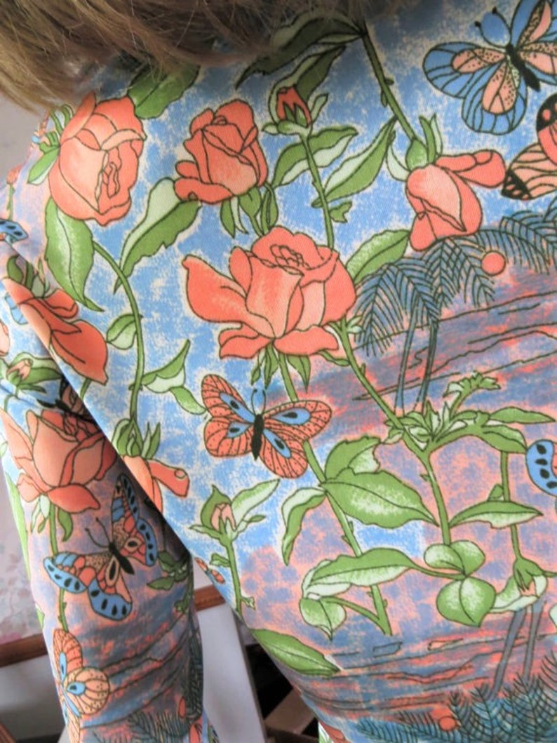 K&ouml;nnte beinhalten: Ein blauer Stoff mit einem Blumenmuster mit rosa Rosen, gr&uuml;nen Bl&auml;ttern und orangefarbenen Schmetterlingen.