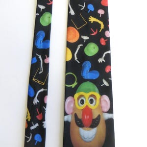 May include: Black novelty tie featuring a cartoon Mr. Potato Head character with a green hat, yellow glasses, and a red nose. The tie has a repeating pattern of colorful shapes and body parts.