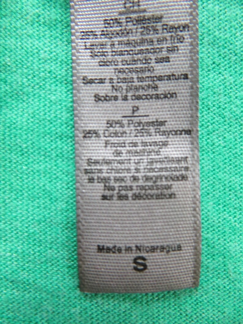May include: A white care label with black text detailing the fabric composition and care instructions for a garment. The label reads "50% Polyester 25% Cotton/25% Rayon" and "Made in Nicaragua". The size "S" is also visible on the label.