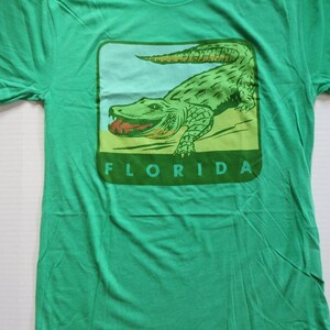 May include: Green t-shirt featuring a vintage-style illustration of an alligator with the word "FLORIDA" printed beneath it.