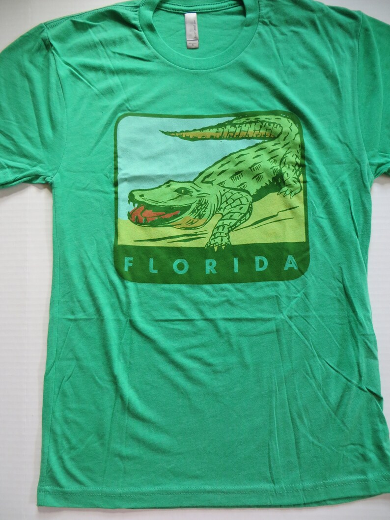 May include: Green t-shirt featuring a vintage-style illustration of an alligator with the word "FLORIDA" printed beneath it.