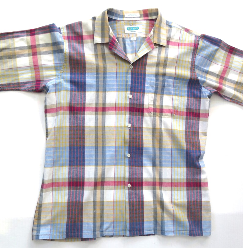May include: A long-sleeved button-down shirt with a plaid pattern in shades of blue, red, yellow, and white. The shirt has a collar and a pocket on the left side.