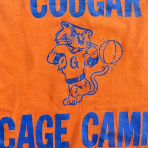 Puede incluir: Camiseta naranja con un gr&aacute;fico azul de un puma de dibujos animados sosteniendo una pelota de baloncesto. El texto "COUGAR CAGE CAMP" est&aacute; impreso en azul en la camiseta.