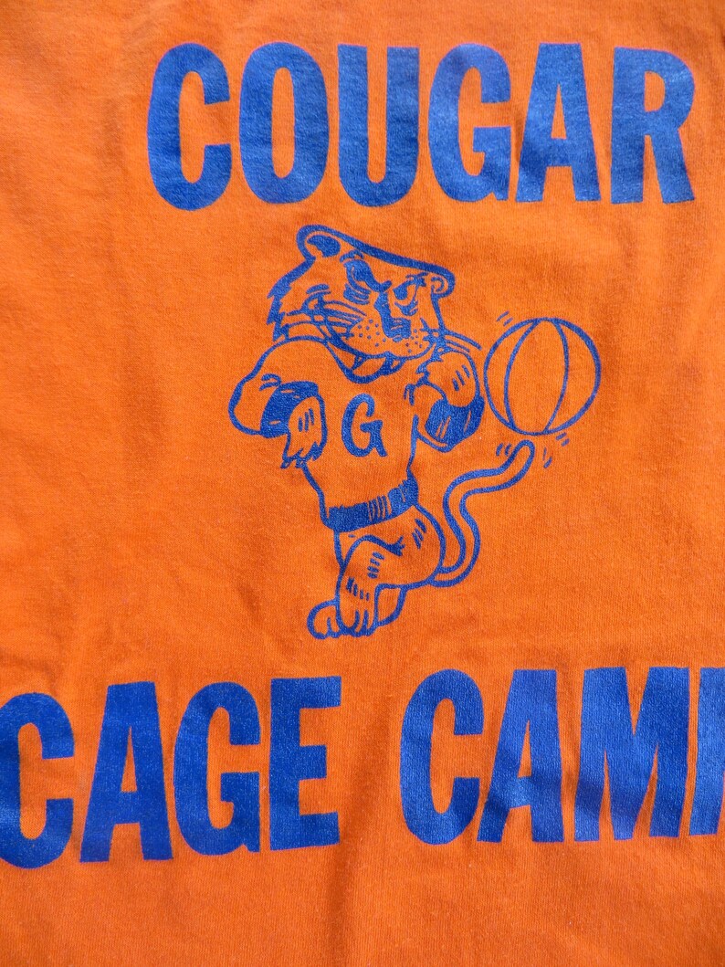 Puede incluir: Camiseta naranja con un gr&aacute;fico azul de un puma de dibujos animados sosteniendo una pelota de baloncesto. El texto "COUGAR CAGE CAMP" est&aacute; impreso en azul en la camiseta.
