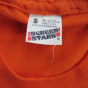 Puede incluir: Camiseta naranja con una etiqueta blanca que dice "S 50% Algod&oacute;n 50% Poli&eacute;ster Hecho en EE. UU. Cuidado en el reverso" y una etiqueta en blanco y negro que dice "Screen Stars".