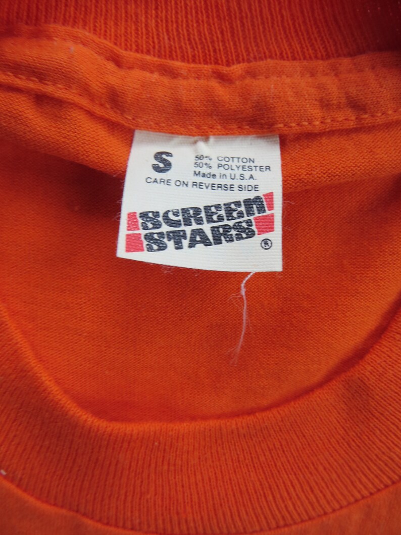 Puede incluir: Camiseta naranja con una etiqueta blanca que dice "S 50% Algod&oacute;n 50% Poli&eacute;ster Hecho en EE. UU. Cuidado en el reverso" y una etiqueta en blanco y negro que dice "Screen Stars".