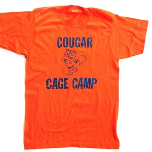 Puede incluir: Camiseta naranja con un gr&aacute;fico azul de un puma de dibujos animados sosteniendo una pelota de baloncesto. El texto "COUGAR CAGE CAMP" est&aacute; impreso en azul.