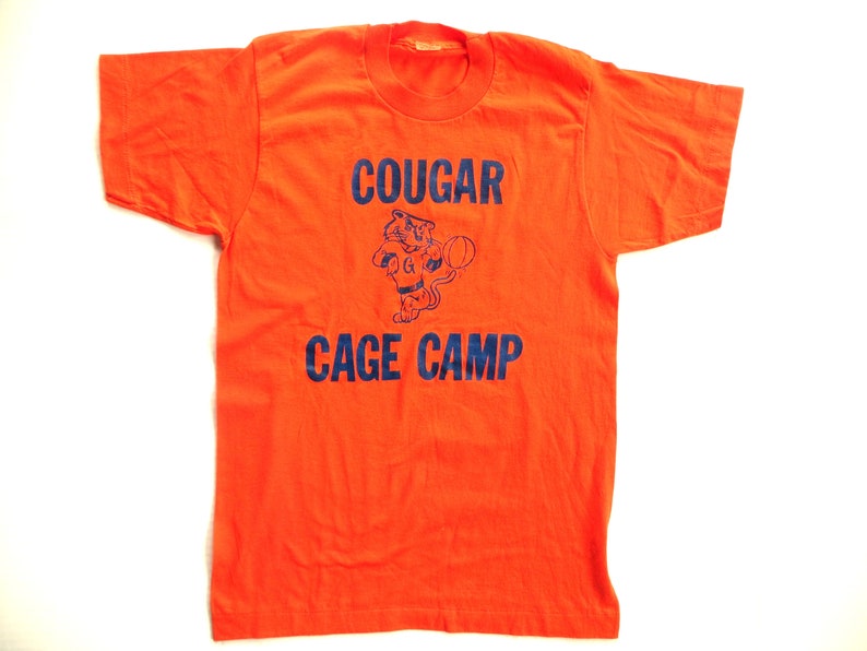 Puede incluir: Camiseta naranja con un gr&aacute;fico azul de un puma de dibujos animados sosteniendo una pelota de baloncesto. El texto "COUGAR CAGE CAMP" est&aacute; impreso en azul.