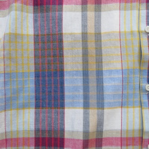 May include: A close-up of a white, blue, yellow, and red plaid fabric. The fabric has a woven texture and is made of cotton.