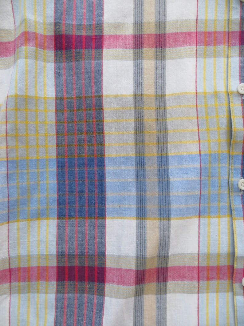 May include: A close-up of a white, blue, yellow, and red plaid fabric. The fabric has a woven texture and is made of cotton.