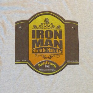 May include: A gray t-shirt with a brown and yellow label that says "Iron Man Miracle Man Ale" with the text "Andy Francis Pick Festivus Brewing Co." and "Sioux City, IA 3.22.2014 FL.OZ."