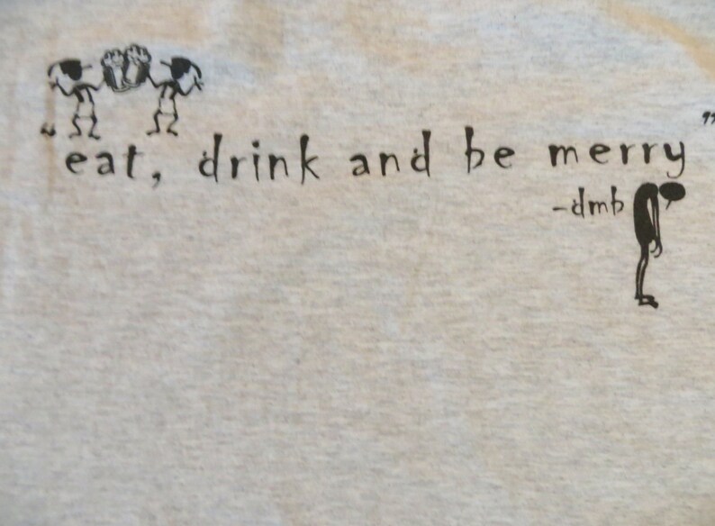 May include: A white t-shirt with black text that reads "eat, drink and be merry" with a small drawing of a person below the text and the words "dmb" written above the person.