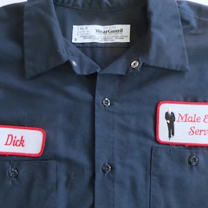 Może przedstawiać: Granatowa koszula robocza z dwiema przednimi kieszeniami i zapięciem na guziki. Posiada dwie haftowane naszywki: jedną z imieniem "Dick" i drugą z napisem "Male Escort Service" oraz małą grafiką mężczyzny w garniturze.