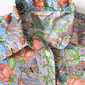 K&ouml;nnte beinhalten: Ein blaues und rosafarbenes Hemd mit Kn&ouml;pfen und einem Blumendruck mit Schmetterlingen und Rosen. Das Hemd hat einen Kragen und ein Etikett mit der Aufschrift "100% Polyester Knit".