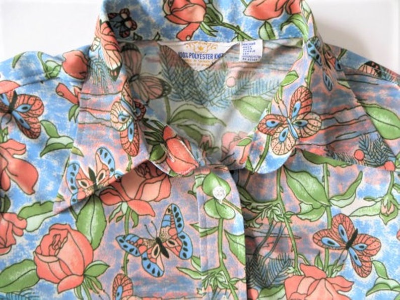 K&ouml;nnte beinhalten: Ein blaues und rosafarbenes Hemd mit Kn&ouml;pfen und einem Blumendruck mit Schmetterlingen und Rosen. Das Hemd hat einen Kragen und ein Etikett mit der Aufschrift "100% Polyester Knit".
