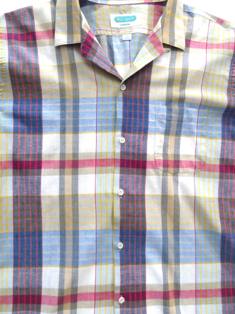 May include: A plaid button-down shirt with a white background and red, blue, yellow, and brown stripes. The shirt has a collar and a pocket on the left side. The label on the shirt reads "Sea Isle by Arrow".