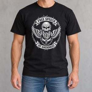 May include: A black short-sleeved t-shirt featuring a white graphic print of a skull wearing a bandana, positioned above a microphone with wings. The design is encircled by barbed wire and includes the text FREE SPEECH and 1ST AMENDMENT.