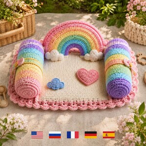 Könnte beinhalten: Eine gehäkelte Spielmatte mit einer cremefarbenen quadratischen Basis und einem gewellten rosa Rand. Sie enthält einen Regenbogenbogen mit weißen Wolken, zwei zylindrische Regenbogen-Seitenteile, eine blaue Wolkenapplikation und ein rosa Herz.