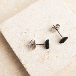 May include: A pair of black gemstone stud earrings with silver posts. The earrings are displayed on a light beige surface.