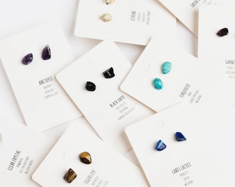 May include: Assortment of small, natural stone earrings displayed on white cards. Each card features the name of the stone and its associated properties. Stones include amethyst, clear crystal, rose quartz, tigers eye, black onyx, lapis lazuli, turquoise, green aventurine, and citrine.