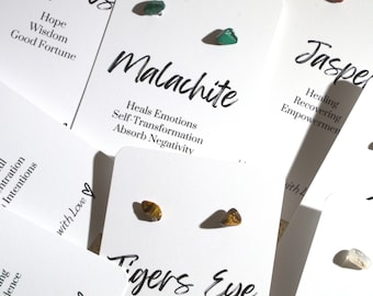 May include: Assortment of small, polished gemstone earrings. Each pair is displayed on a white card with the name of the stone and its associated properties. Stones include turquoise, malachite, jasper, tigers eye, and citrine.