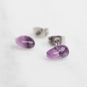 May include: A pair of silver stud earrings with purple amethyst stones. The stones are oval shaped and have a natural, irregular look.
