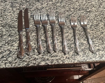 Peut inclure: Un ensemble de huit couverts en acier inoxydable argenté comprenant deux couteaux de table et six fourchettes. Chaque pièce présente un motif ornemental en relief sur le manche, avec une finition polie et un design classique effilé.