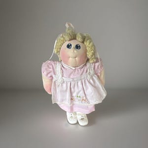 Könnte beinhalten: Eine Stoffpuppe mit blondem Wollhaar, blauen Augen und rosigen Wangen. Die Puppe trägt ein hellrosa Kleid mit einer weißen Schürze, die mit gestickten Reh- und Kleemotiven verziert ist, dazu weiße Schuhe und ein weißes Haarband im Haar.
