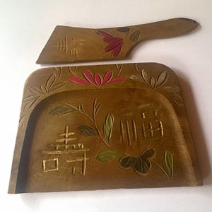 Könnte beinhalten: Ein Kehrblech- und Handfeger-Set aus Holz mit geschnitzten Blumenmotiven und chinesischen Schriftzeichen. Das Holz hat einen mittelbraunen Ton mit handbemalten Akzenten in Pink, Grün und Gold. Das Kehrblech hat eine breite, flache Basis.