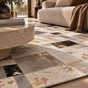 May include: A rectangular area rug featuring a patchwork design of square and rectangular tiles in shades of cream, gray, and black. The surface displays a textured finish with metallic gold accents scattered across the dark and light panels.