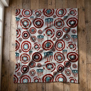 Op de afbeelding: Een witte fleece deken met een herhalend patroon van rode, witte en blauwe cirkelvormige schilden met een centrale ster, afgewisseld met blauwe schildvormige emblemen met verticale rode strepen aan de onderkant, in een verweerd grafisch ontwerp.