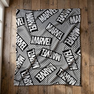 Può includere: Una coperta rettangolare con un motivo ripetitivo in bianco e nero in stile fumetto. Il design presenta la parola MARVEL stampata in grassetto bianco maiuscolo all'interno di riquadri rettangolari neri su uno sfondo a pois bianchi.