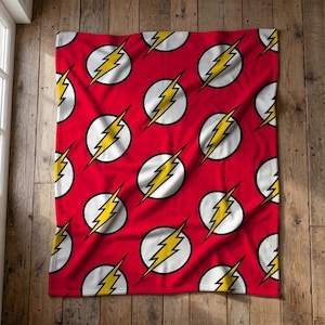 Peut inclure: Une couverture polaire rouge avec un motif répétitif de cercles blancs, chacun contenant un emblème d'éclair jaune. Le tissu présente une texture douce et est étalé à plat, affichant le design graphique cohérent sur toute la surface.