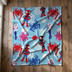 Könnte beinhalten: Eine hellblaue Fleecedecke mit einem sich wiederholenden Muster aus Zeichentrickfiguren wie Spider-Man, Spider-Gwen und Miles Morales, durchsetzt mit roten und blauen Spinnensymbolen, weißen Spinnennetzen und kleinen weißen Wolkenformen.