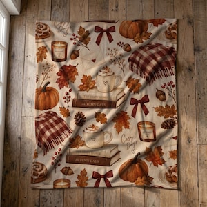 May include: A cream-colored throw blanket featuring an autumn-themed pattern of orange pumpkins, brown fall leaves, red bows, plaid blankets, cinnamon rolls, and mugs of hot cocoa resting on books labeled The Cozy Home and Autumn Days.