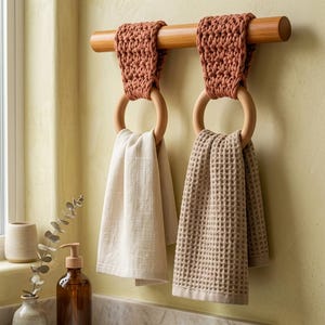 Op de afbeelding: Twee terracotta-kleurige gehaakte lussen hangen aan een horizontale houten stang, elk ondersteund door een gladde houten ring. Een effen crèmekleurige handdoek hangt aan de linkerring, terwijl een beige handdoek met wafelstructuur rechts hangt.