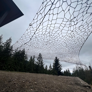 Könnte beinhalten: Ein großes schwarzes Seilnetz mit unregelmäßigem, spinnennetzartigem Muster, das in der Luft hängt. Die Struktur besteht aus dicken, geknoteten Seilsegmenten, die verschiedene polygonale Formen bilden, über einem Kiesboden und einem Nadelwald.