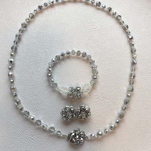 Peut inclure: Un ensemble de bijoux composé d'un collier, d'un bracelet et d'une paire de boucles d'oreilles. Chaque pièce est fabriquée à partir de perles en cristal transparent et argenté à facettes. Le collier et le bracelet présentent une fleur centrale.