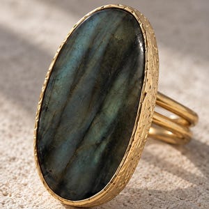 May include: A gold ring featuring a large, oval-shaped labradorite gemstone with dark blue and grey tones. The stone is set in a textured gold bezel, and the ring band consists of two thin, smooth gold wires that connect to the setting.