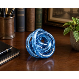 May include: A decorative sculpture shaped like a complex, continuous knot made of translucent, vibrant blue glass. The object features smooth, rounded surfaces that catch the light, creating a series of interconnected loops and curves in a circular form.