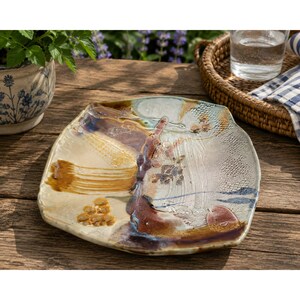 May include: A square ceramic plate with rounded corners, featuring a glazed surface with abstract brushstrokes in shades of cream, brown, and blue, alongside textured honeycomb patterns and smooth, glossy areas across the centre and edges.