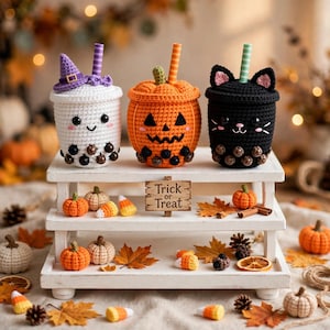 Könnte beinhalten: Drei gehäkelte Boba-Teebecher, bestehend aus einem weißen Geist mit violettem Hut, einem orangefarbenen Kürbis mit Jack-o'-Lantern-Gesicht und einer schwarzen Katze. Jeder Becher hat einen Strohhalm und dunkle Perlen am Boden auf einem Holzständer.