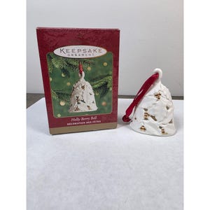 May include: A white ceramic bell ornament featuring a cut-out holly leaf pattern with gold-colored berries. The bell is attached to a red ribbon for hanging and is displayed next to its original rectangular box labeled Keepsake Ornament Holly Berry Bell.